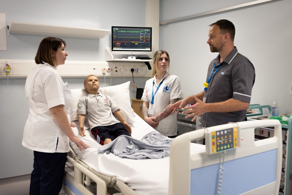 Clinical education team with student nurse standing around a hospital bed with monitoring equipment and dummy patient in bed